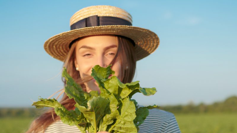 Young woman with hat holds sugar beet in front of her face.