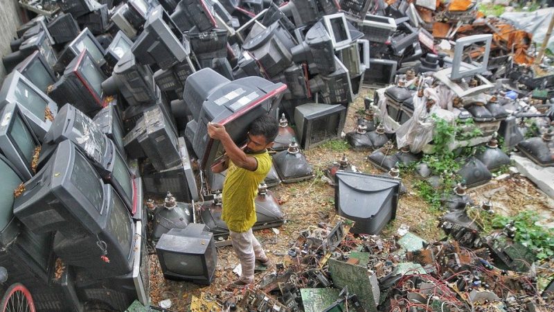 Man at Bangladesh garbage dump drags broken monitor on his shoulders.